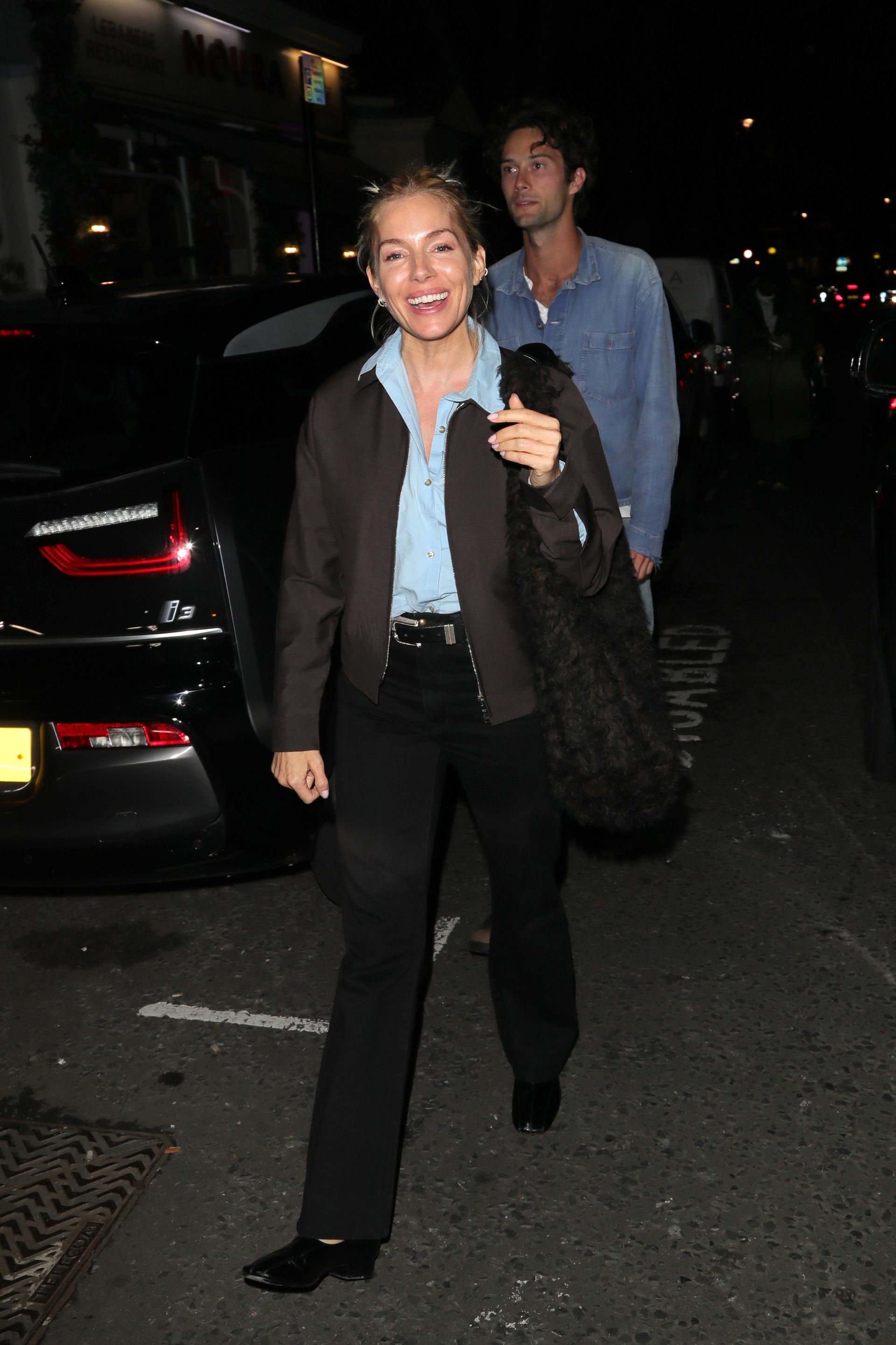 Sienna Miller steps out in London wearing black jeans with square-toe flats, a blue shirt and a dark brown jacket.