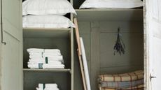 A pale green linen closet with pillows in white pillowcases, folded white sheets, folded gingham blankets, an ironing board, and a sprig of lavender hung up with a blue ribbon.