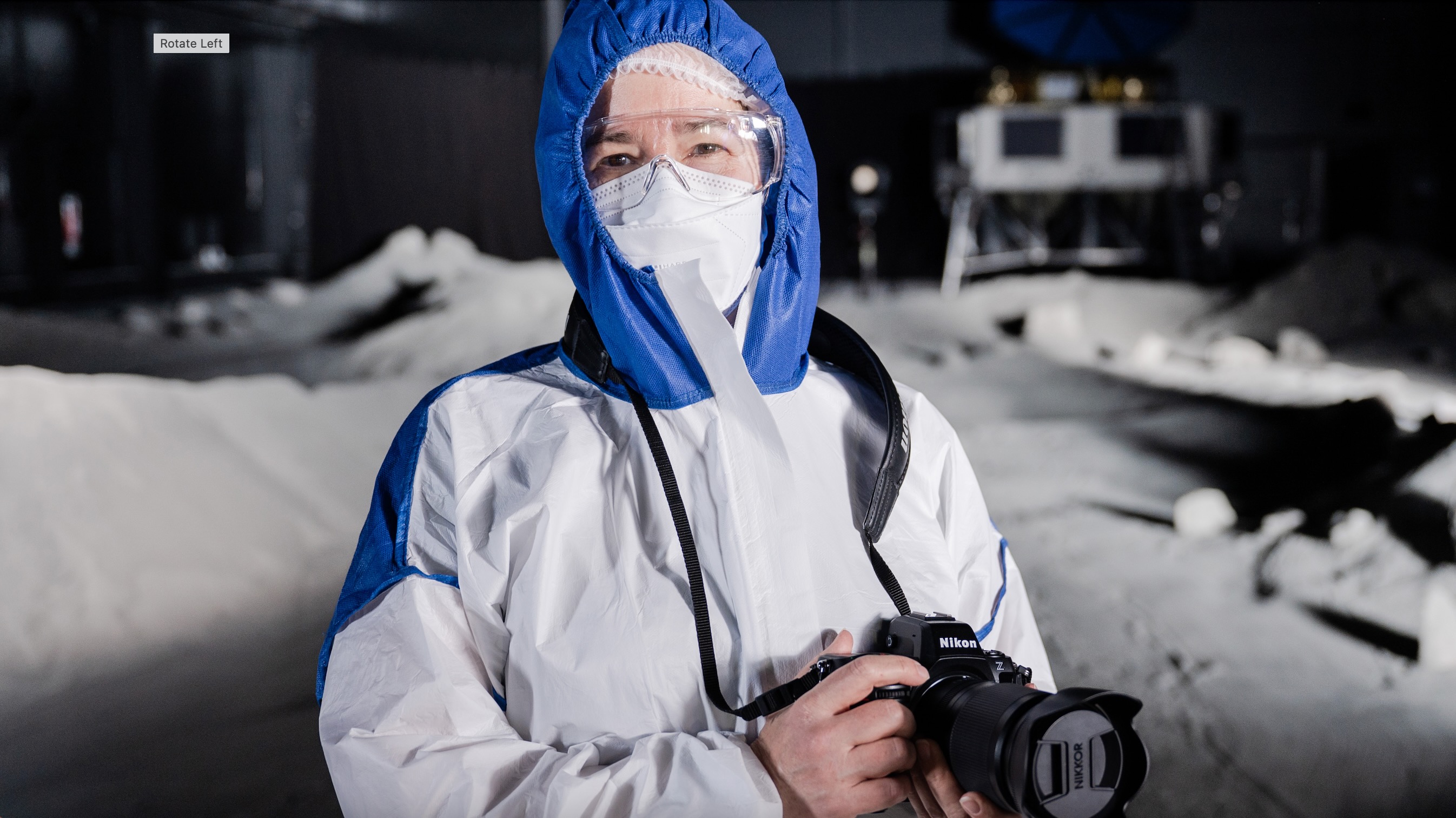 closeup of a woman in a simulated lunar landscape wearing white protective gear and holding a black camera