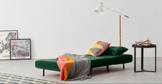 Made.com green Haru sofa bed in a white minimalist room, with framed artwork resting against the wall, a white and wood floor lamp, and a black side table