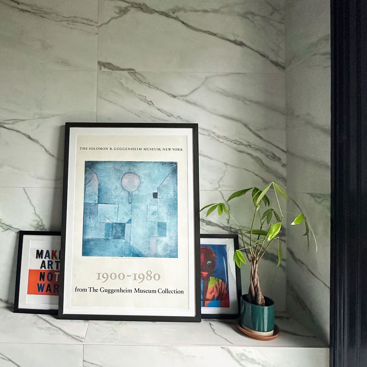Pot plant and three framed artworks on ledge in marble bathroom