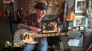 Kelly Garni poses with his blue bass guitar and a picture of the early line-up of Quiet Riot 