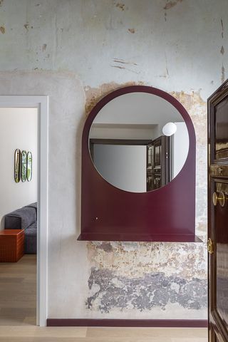 The rustic interiors of a modern apartment feature a cherry-shaded wall mirror, a grey couch and three paintings placed next to each other.