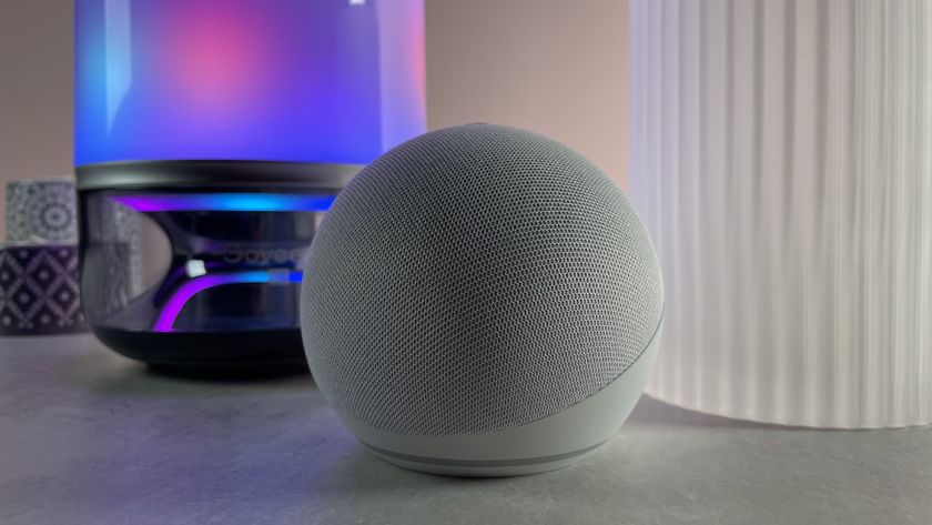 The Echo Dot is pictured in a dim room in front of a large lamp that&#039;s lit up in pretty colors.