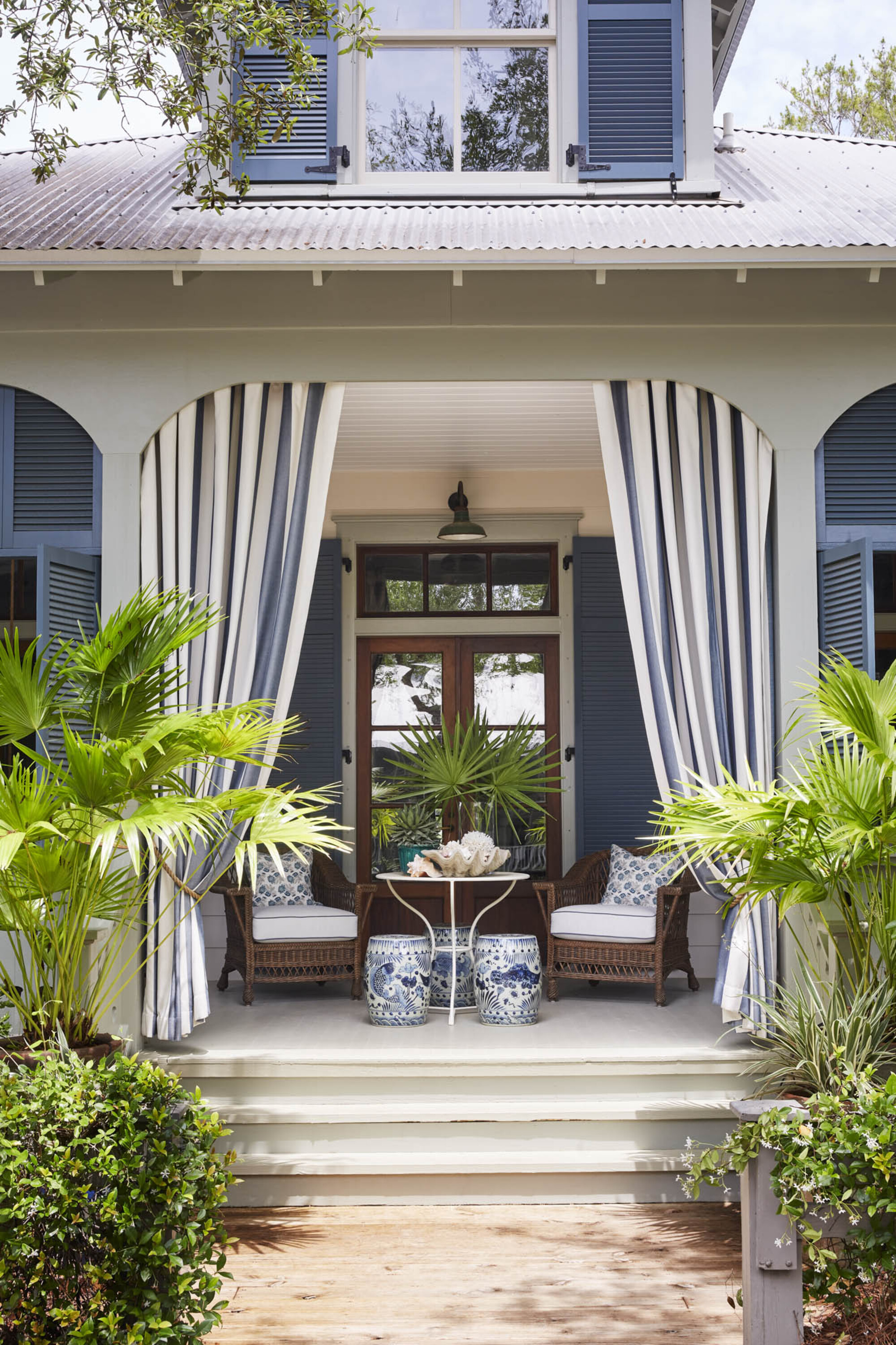 A porch with garden furniture, blue walls, blue shutters, and blue and white striped curtains draped within an arch