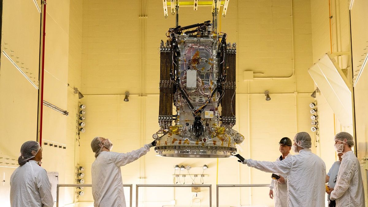 Five people wearing white protective equipment stand around a rectangular spacecraft hanging from the ceiling of a white-walled clean room