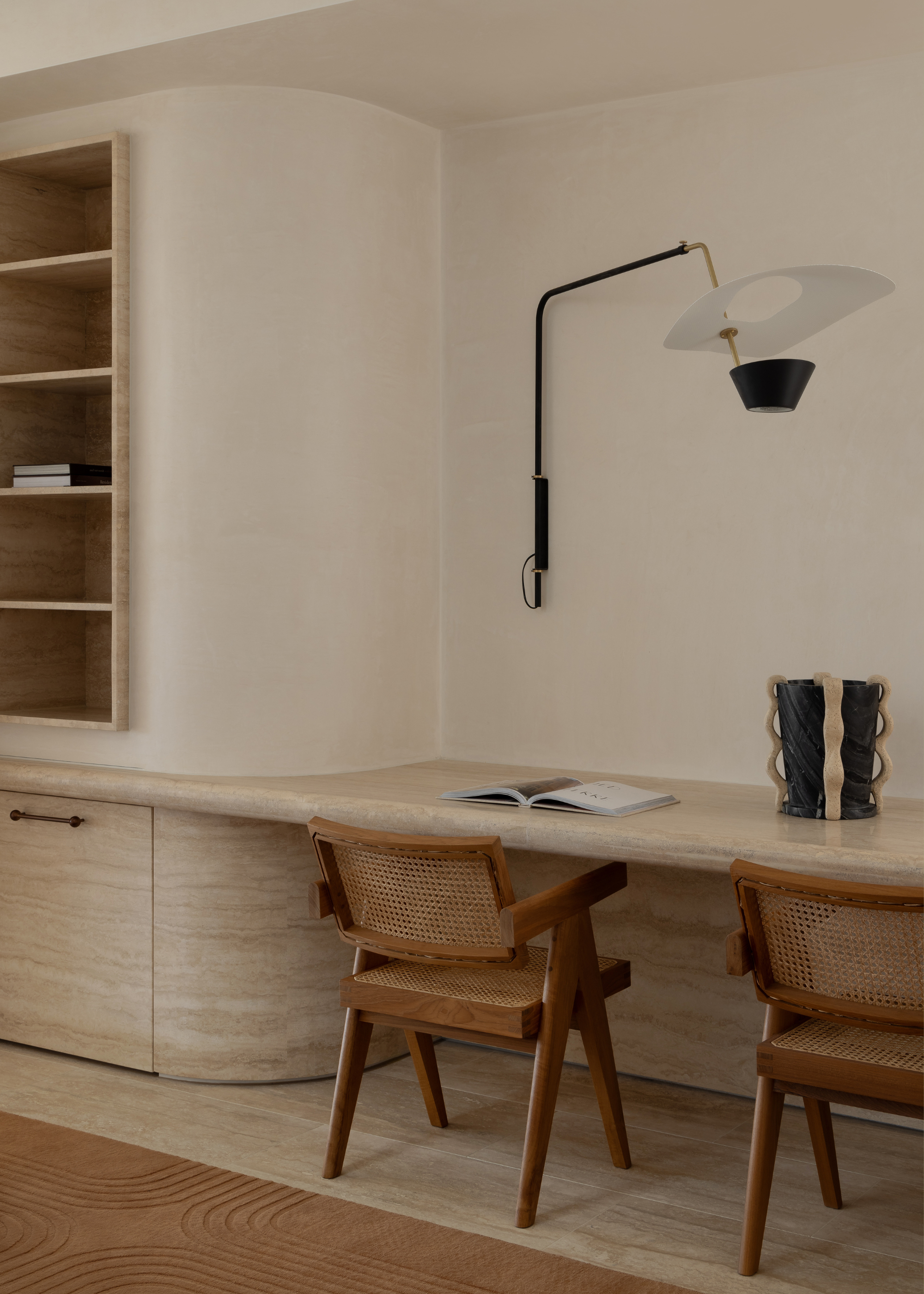 A study space with a pair of rattan and wood chairs, a marble table, a wall-mounted lamp light, an open book and a structural vase