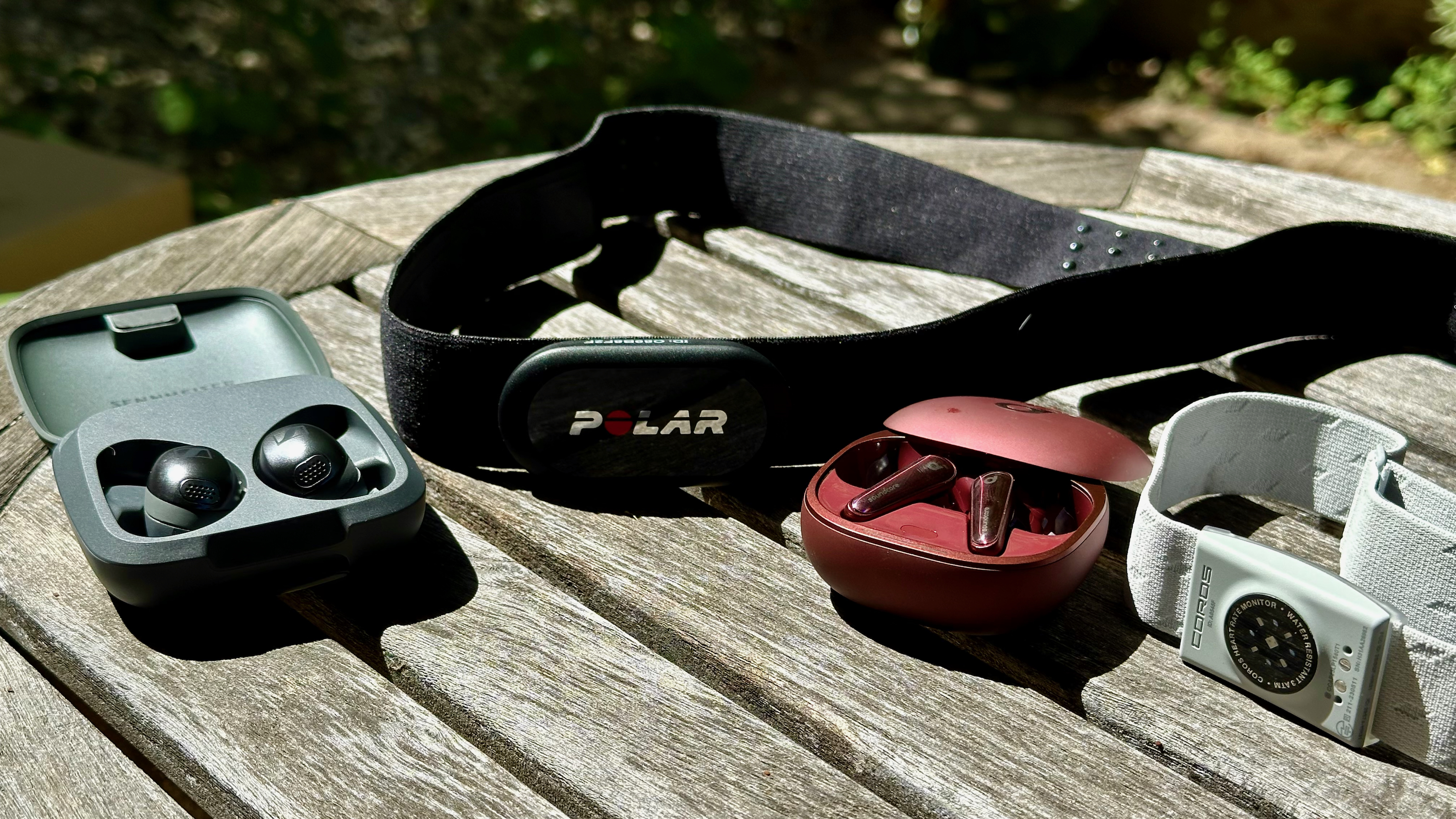 The Polar H10 and COROS Heart Rate Monitor sitting on a table next to two heart rate earbuds, the Sennheiser Momentum Sport and Anker Soundcore Liberty 4.