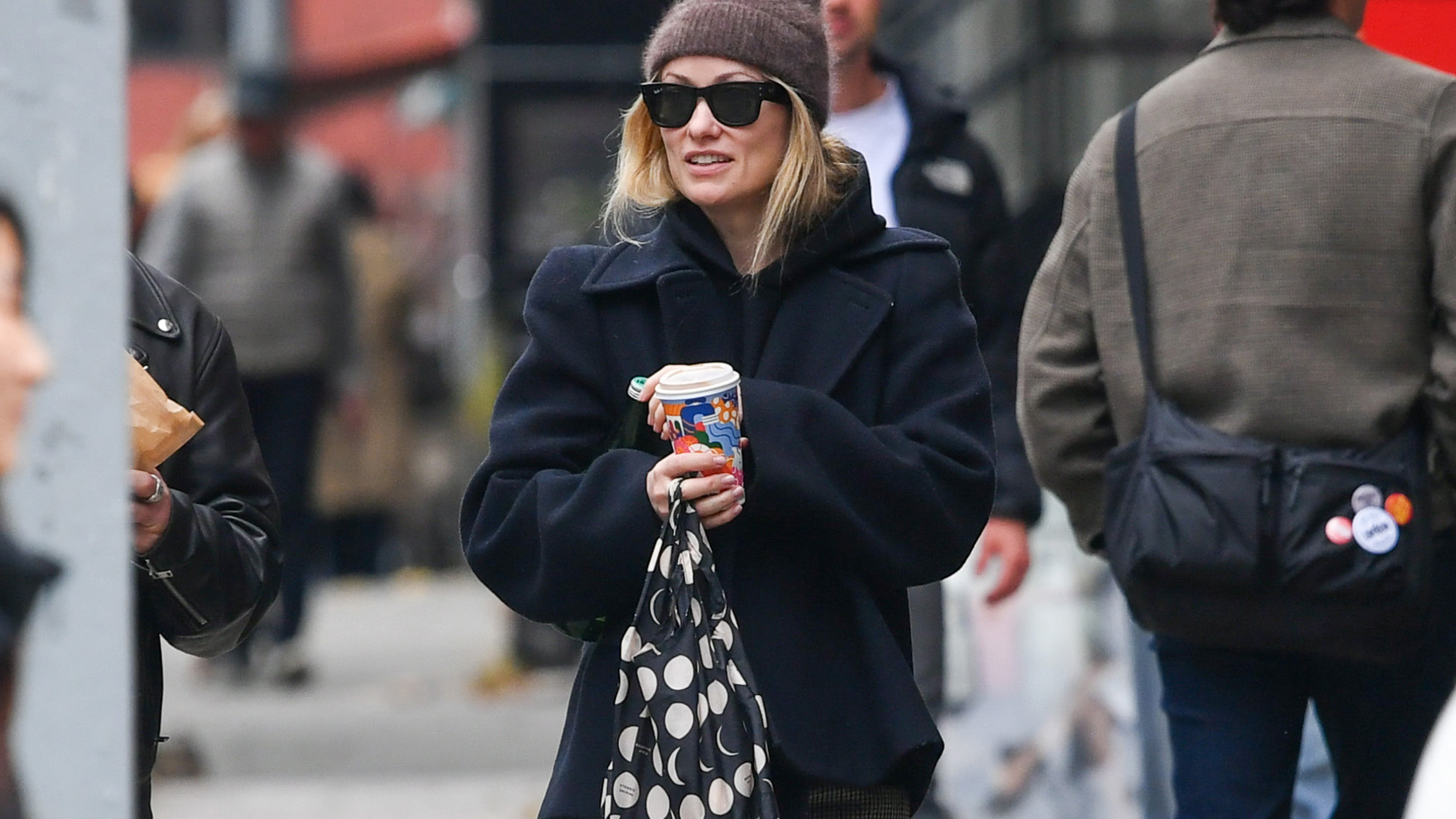 11/25/2025: Olivia Wilde is pictured on a stroll with a male companion in New York City. The American actress wore a wool beanie, black coat, checkered trousers, and Adidas trainers.