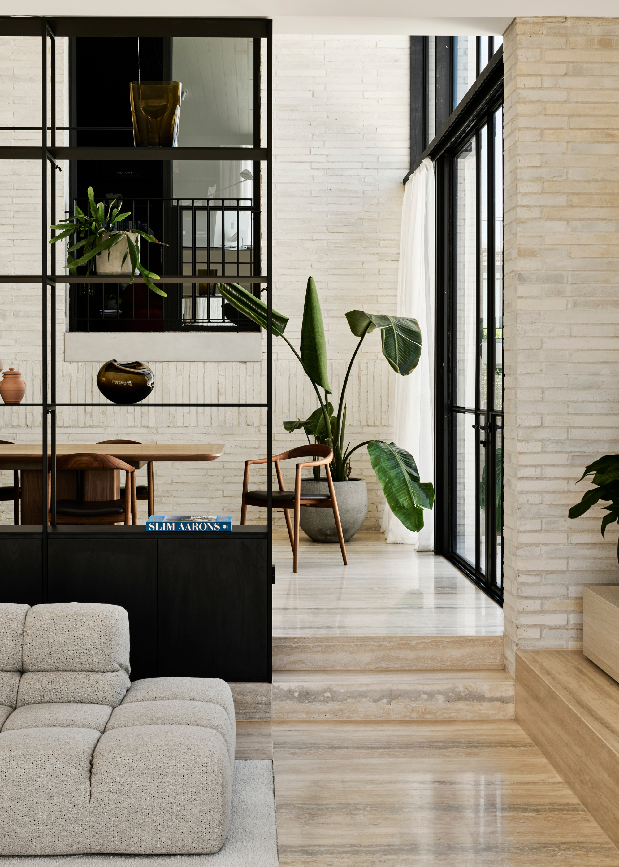 A bright living room with marble floors, a gray-white couch, a black shelving case with plants, books, and vases, and a potted houseplant by a wood dining set