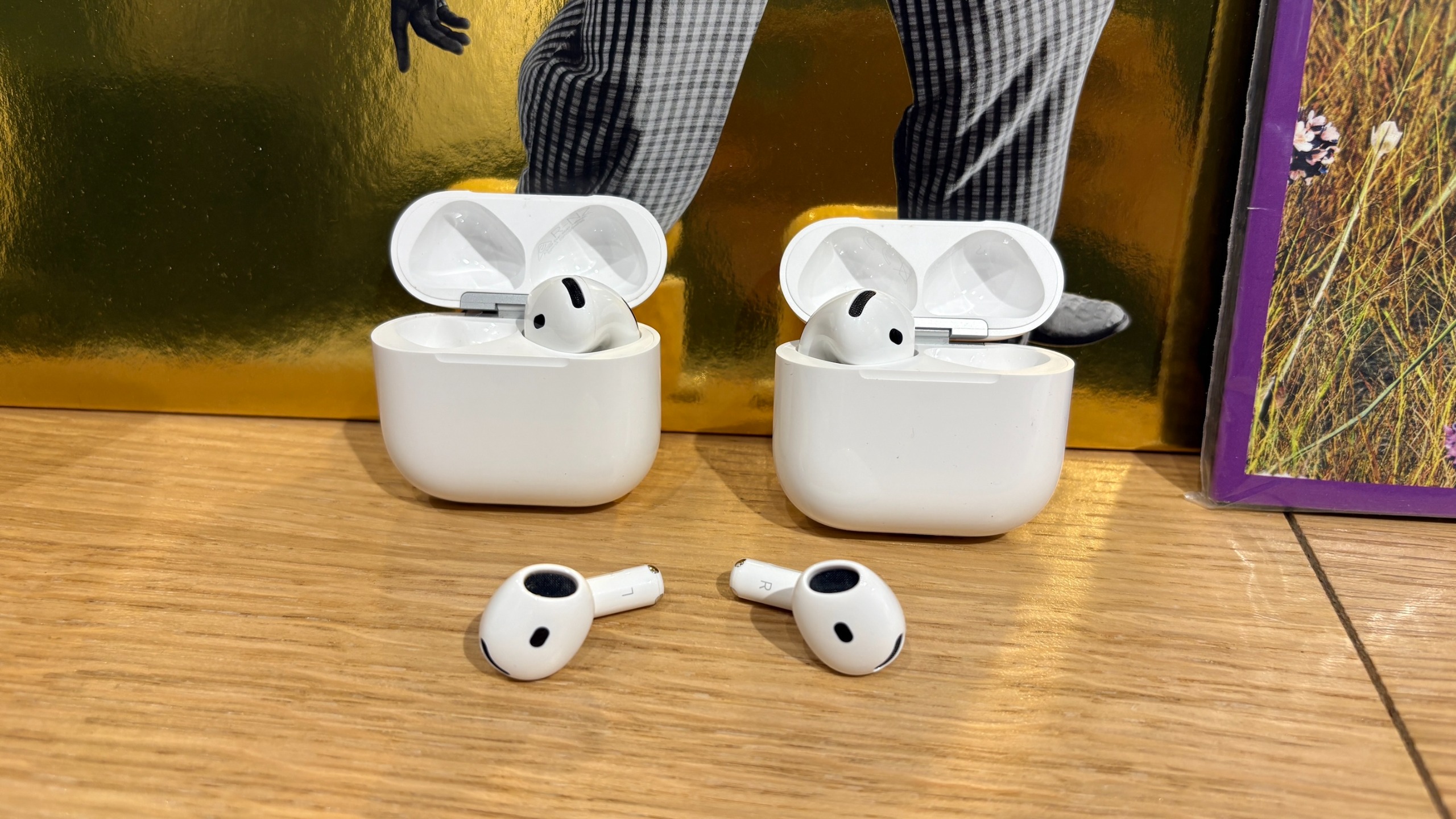 The AirPods 5 and AirPods 4 next to each other on a wooden surface, with one earbud each in their respective charging cases and one lying in front of them.
