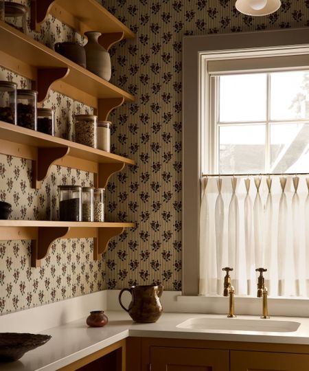 a sweet home pantry with ochre yellow cabinetry and shelving, small sink, window with cafe curtain and floral ditsy wallpaper