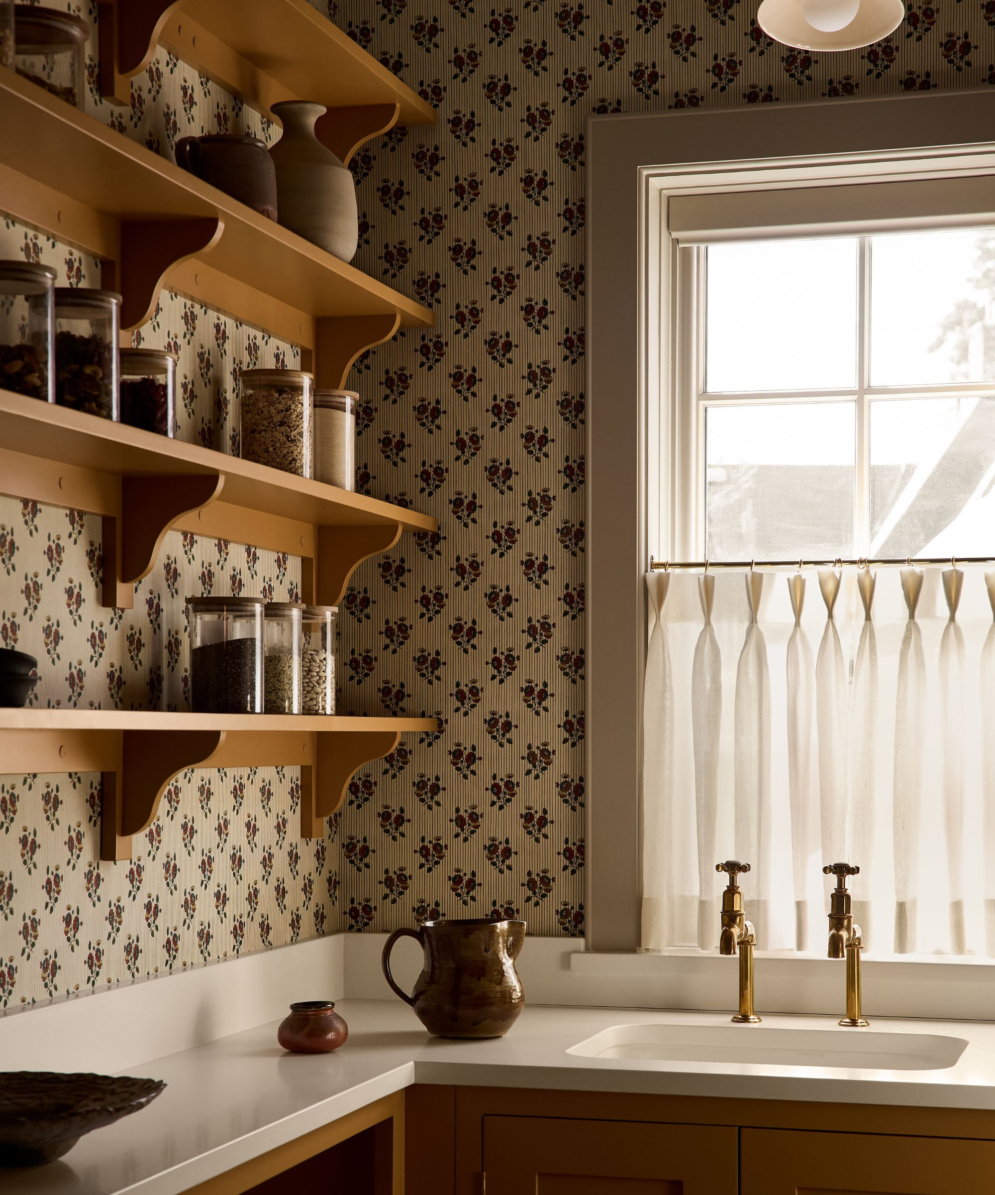 a sweet home pantry with ochre yellow cabinetry and shelving, small sink, window with cafe curtain and floral ditsy wallpaper