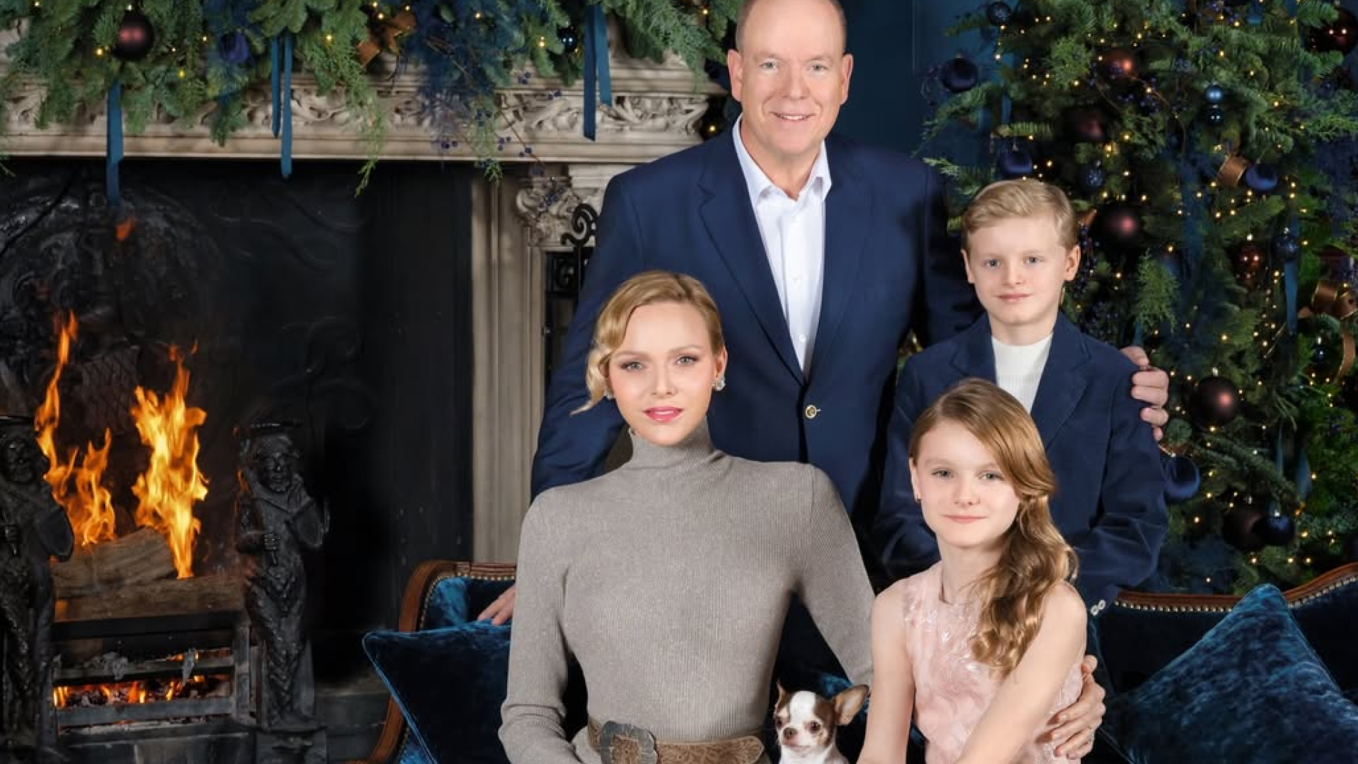 Princess Charlene wears a long taupe dress with a pleated skirt, long sleeves, and a turtle neck, and poses next to a burning fireplace and a Christmas tree with her pet dog, her twin children, Princess Gabriella and Prince Jacques, and husband Prince Albert 