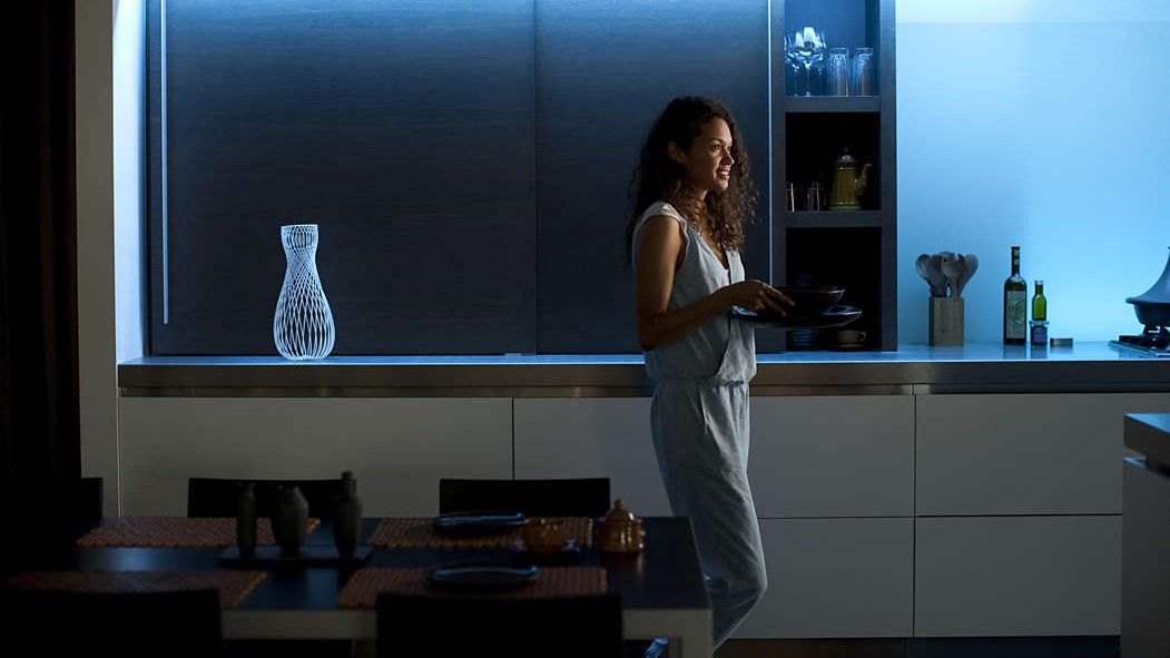 Philips Hue Lightstrip deals 2023, image shows woman in room with blue uplighting on the shelves behind her