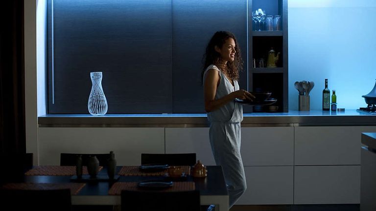 Philips Hue Lightstrip deals 2022, image shows woman in room with blue uplighting on the shelves behind her