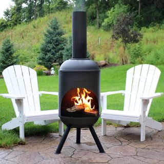 a tall black chiminea on an outdoor patio in the middle of two white deck chairs, with grass beyond