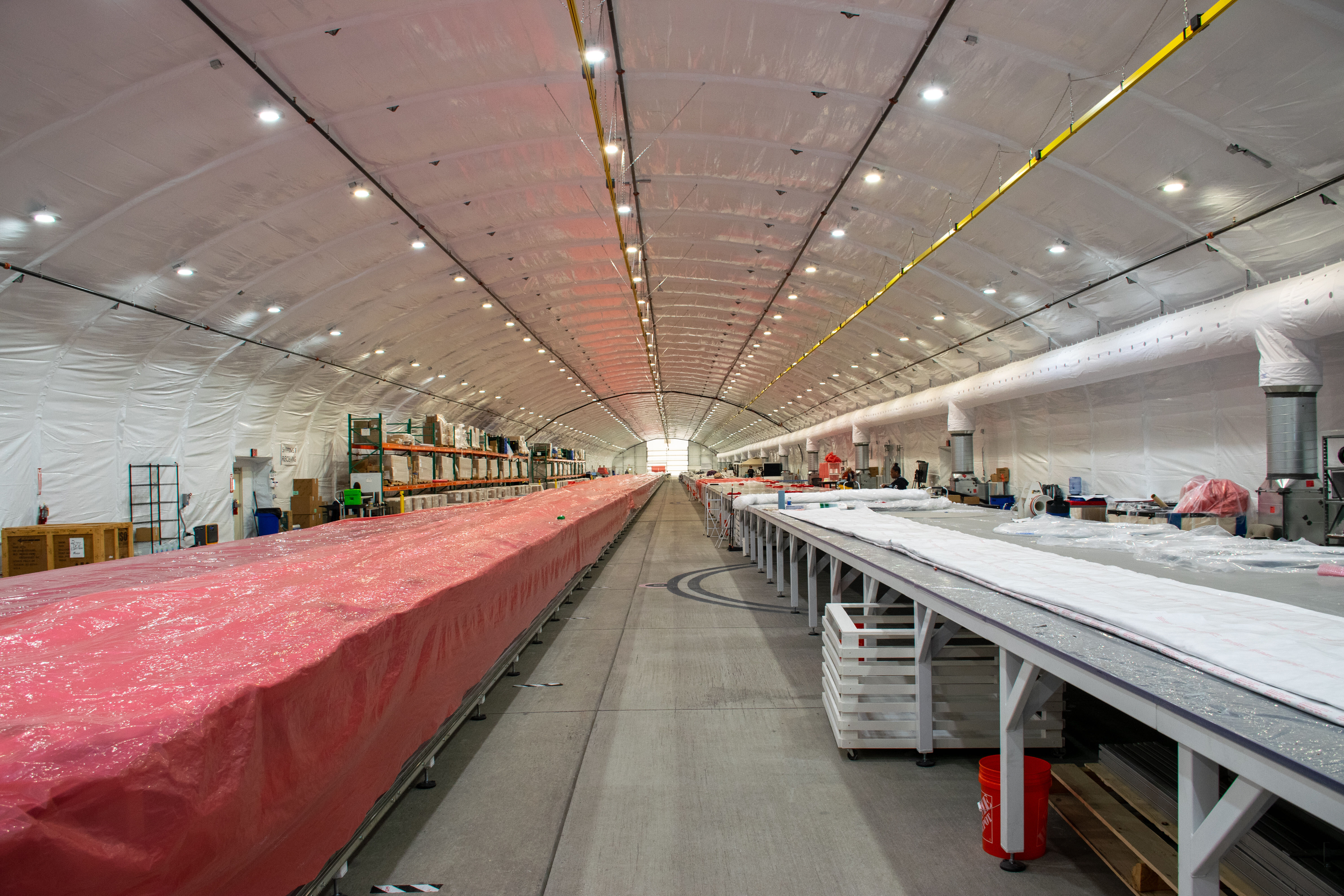 two endlessly long tables stand parallel in a long white hanger. the left table is draped in a red tarp.