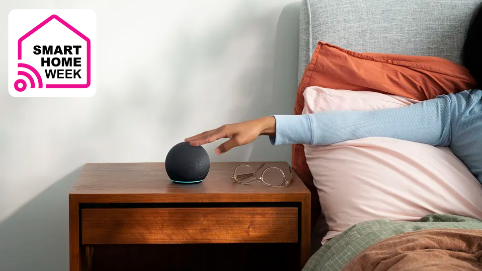 Amazon Echo Dot on a table next to a person in bed with their arm outstretched to turn off its alarm, as well as a superimposed label reading &#039;Smart Home Week&#039;