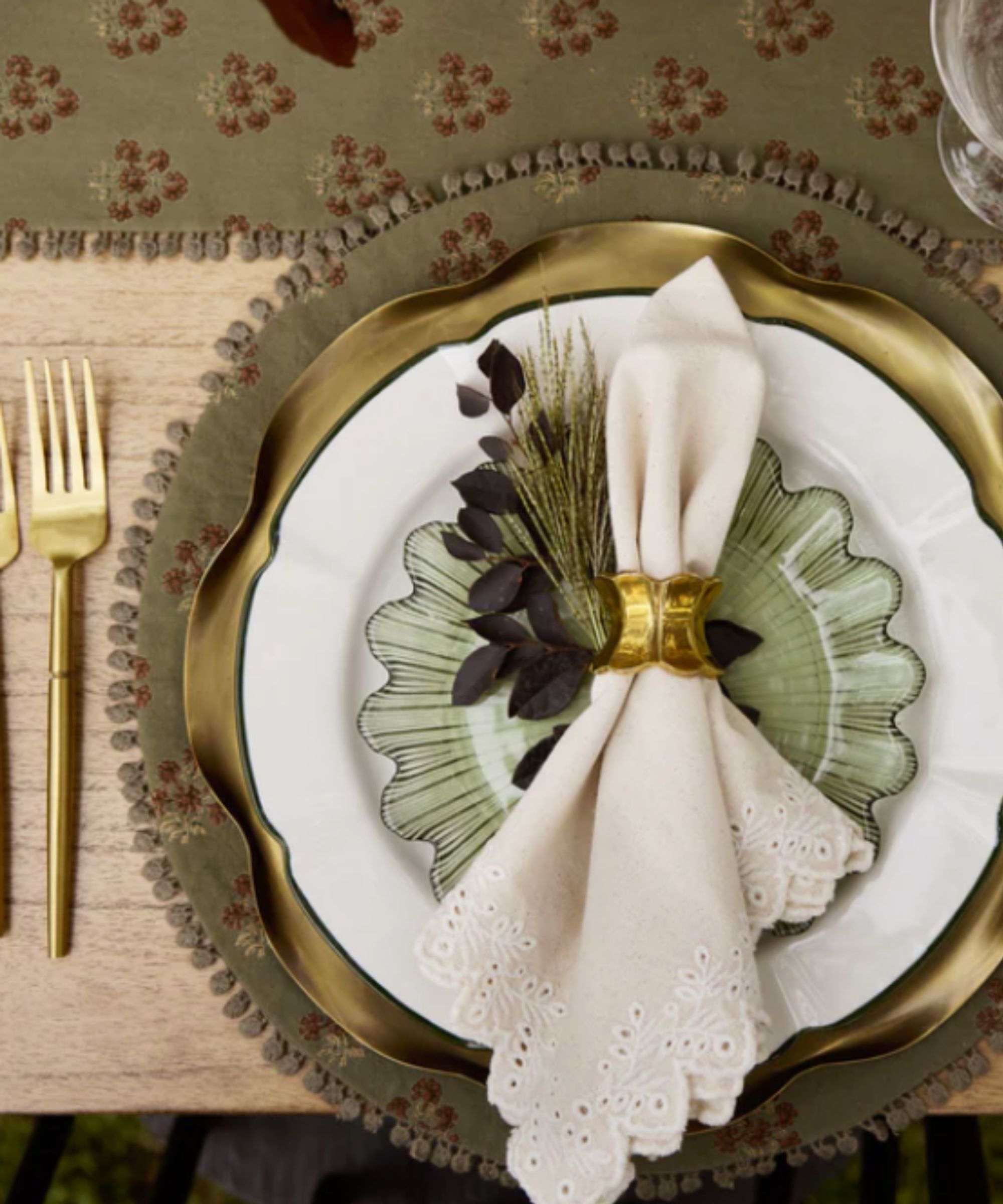 Wooden table with olive green floral place mats, gold charger plates and white crockery