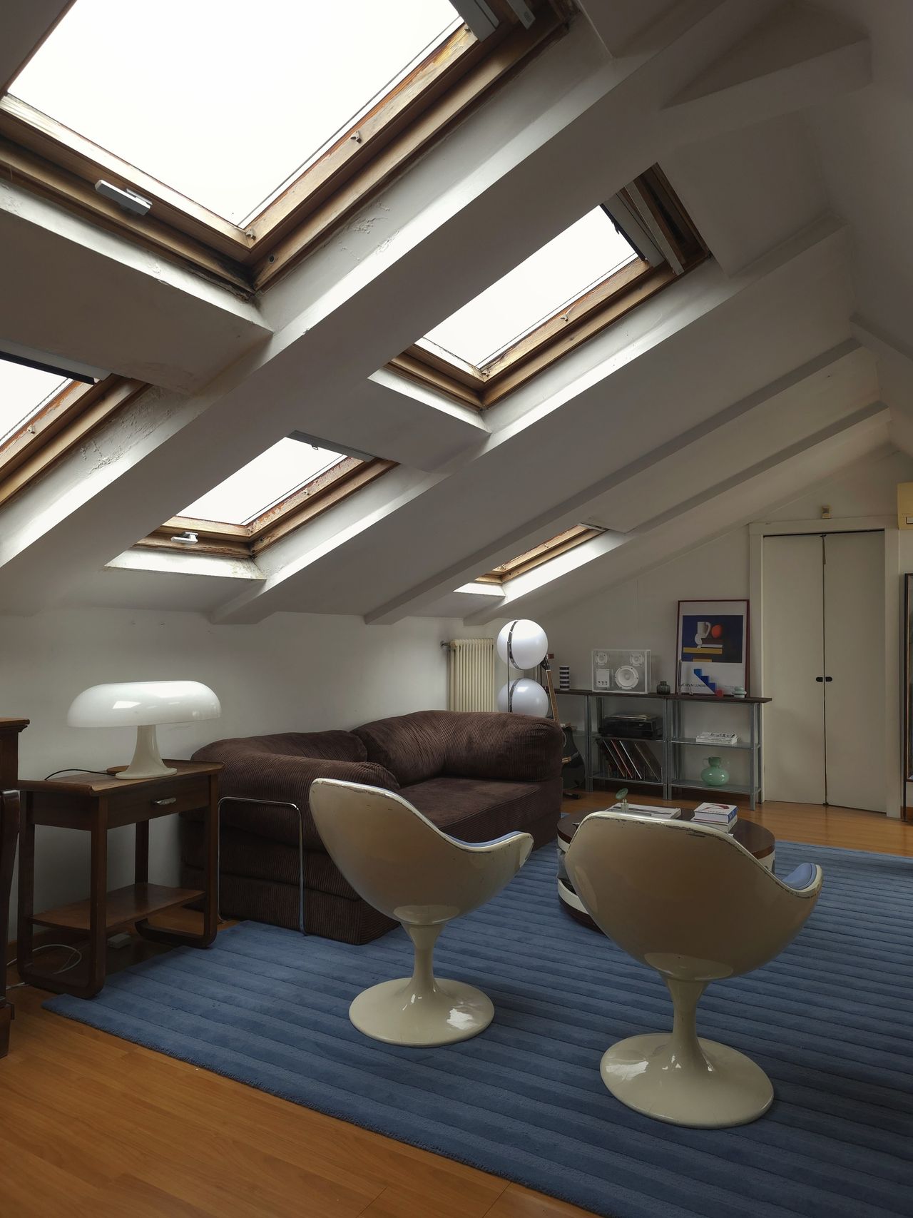 A mid-century modern-style living room with white slanted ceilings with skylights in them. There is a bug rug, a dark brown sofa, two blue and cream egg chairs, and a white wooden coffee table.