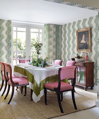 Dining room with walls papered in bold green foliage pattern, matching curtains, table laid for dinner and pink upholstered dining chairs.