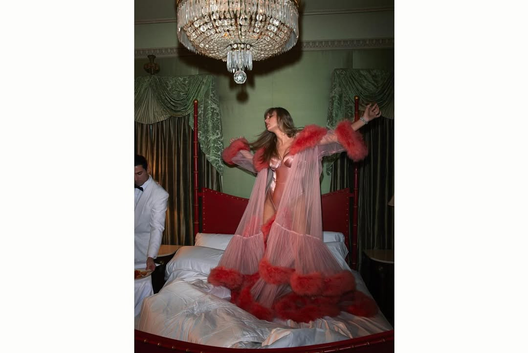 Taylor Swift on a bed in a hotel wearing a feathered robe with satin Fleur du Mal underwear