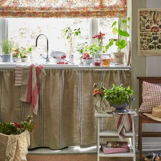 A kitchen with herbs and fresh vegetables on the counter and under-sink curtain cover