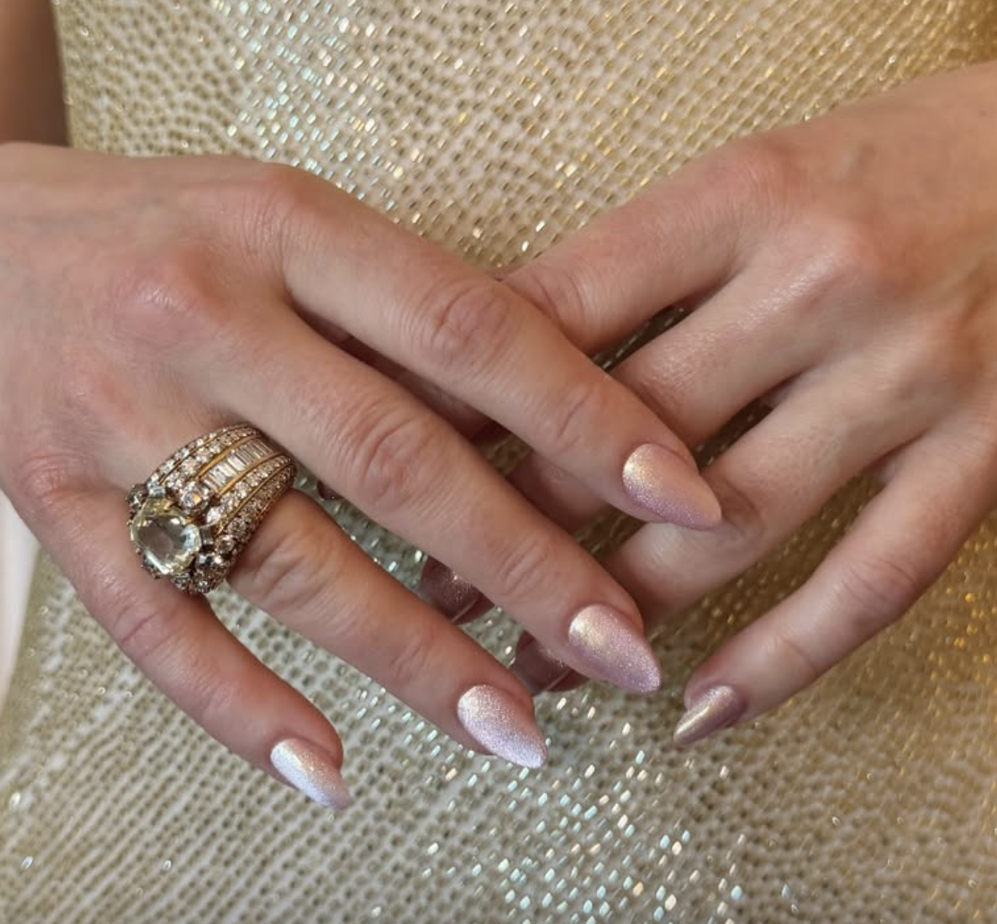 a close up of a woman's hands wearing a gold ring with a metallic baby pink manicure