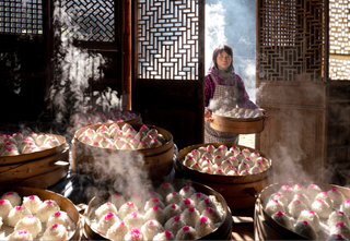 Older woman carrying tray of steaming red paste balls through a wooden door into a room with similar trays piled on a table