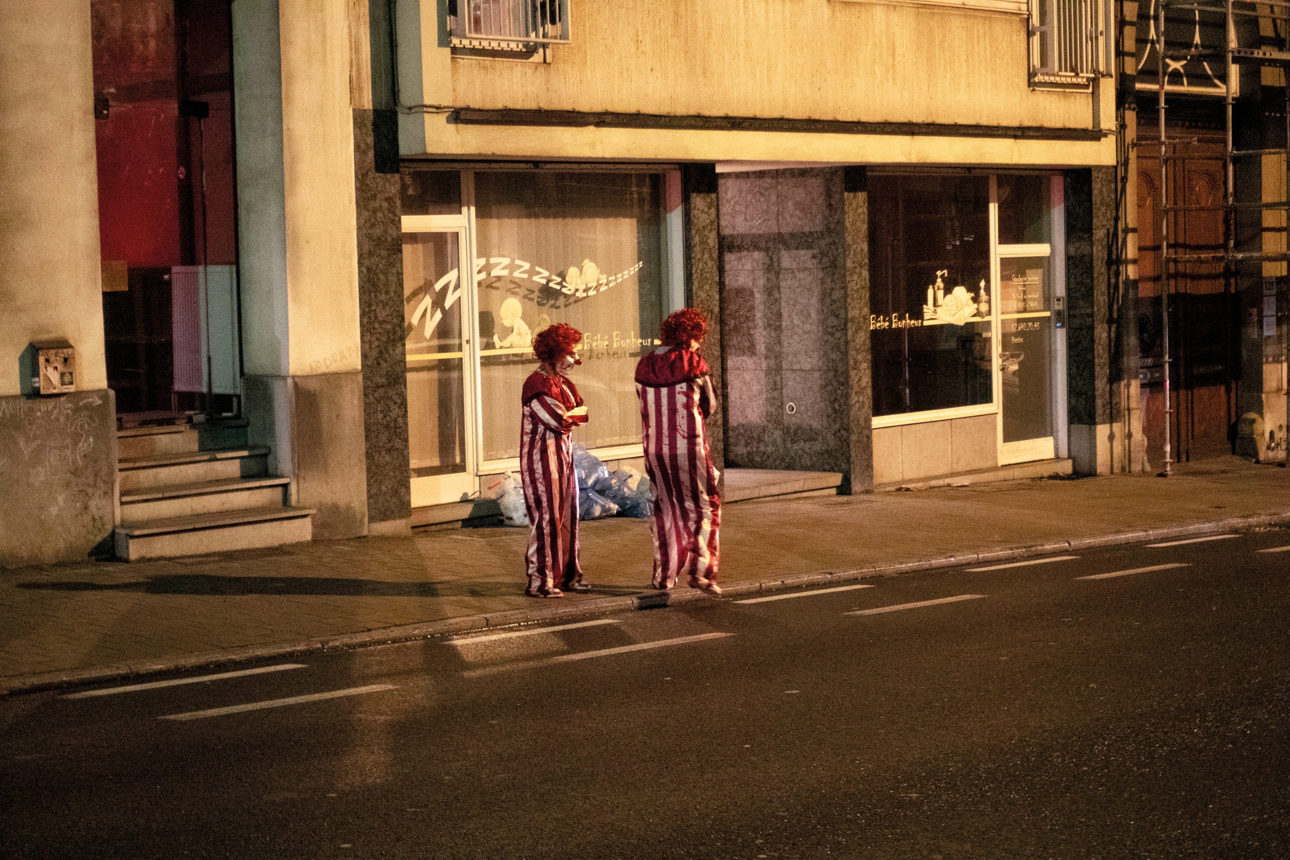 Two clowns stand talking on a night-time pavement outside a shuttered baby goods shop, lit by the shop's glowing window display.