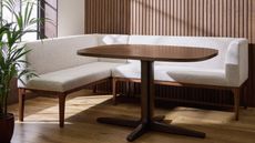 A white upholstered corner dining bench with a wooden frame in front of a wood paneled wall with a dining table in the center and next to a window and potted houseplant