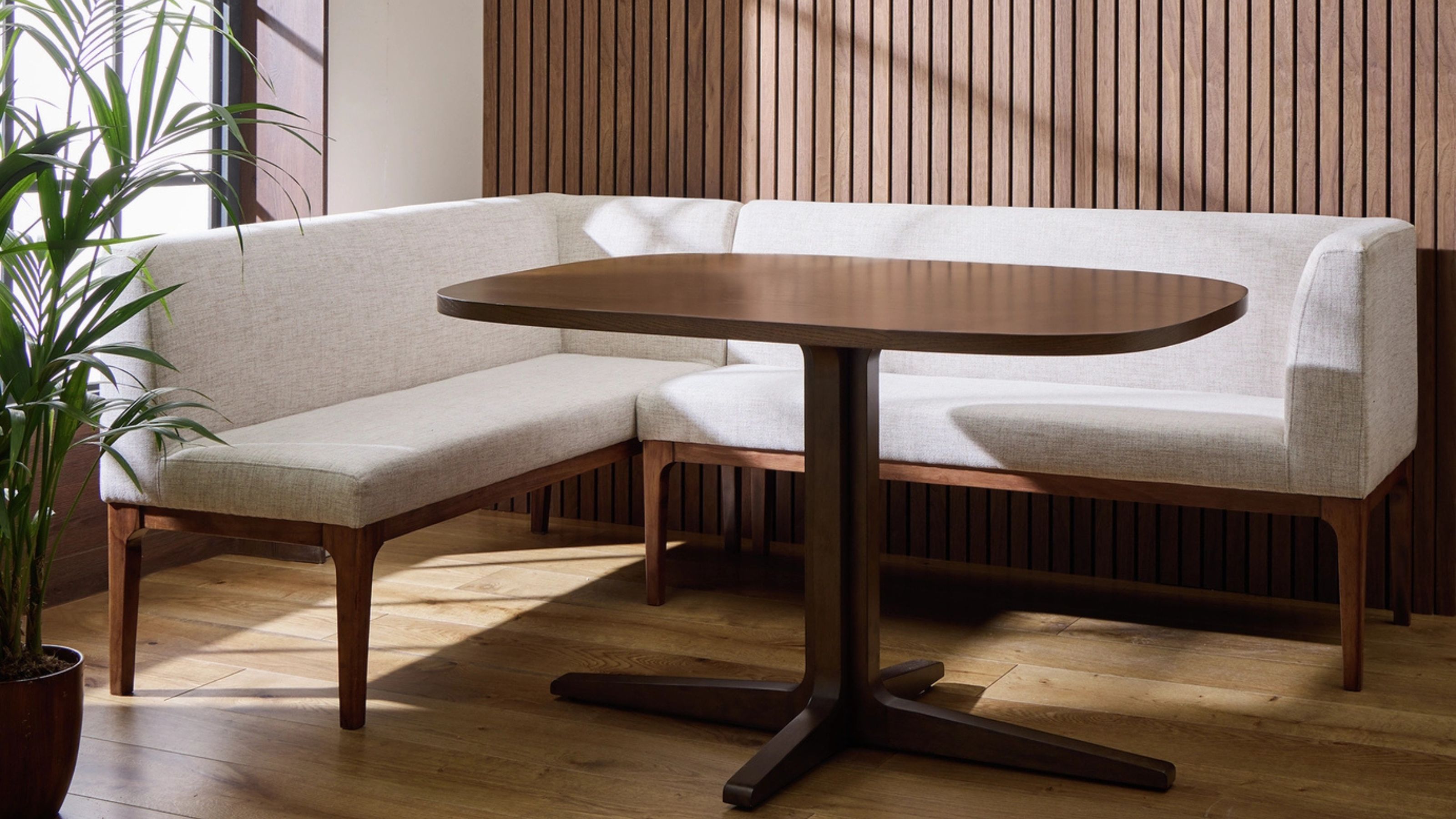 A white upholstered corner dining bench with a wooden frame in front of a wood paneled wall with a dining table in the center and next to a window and potted houseplant