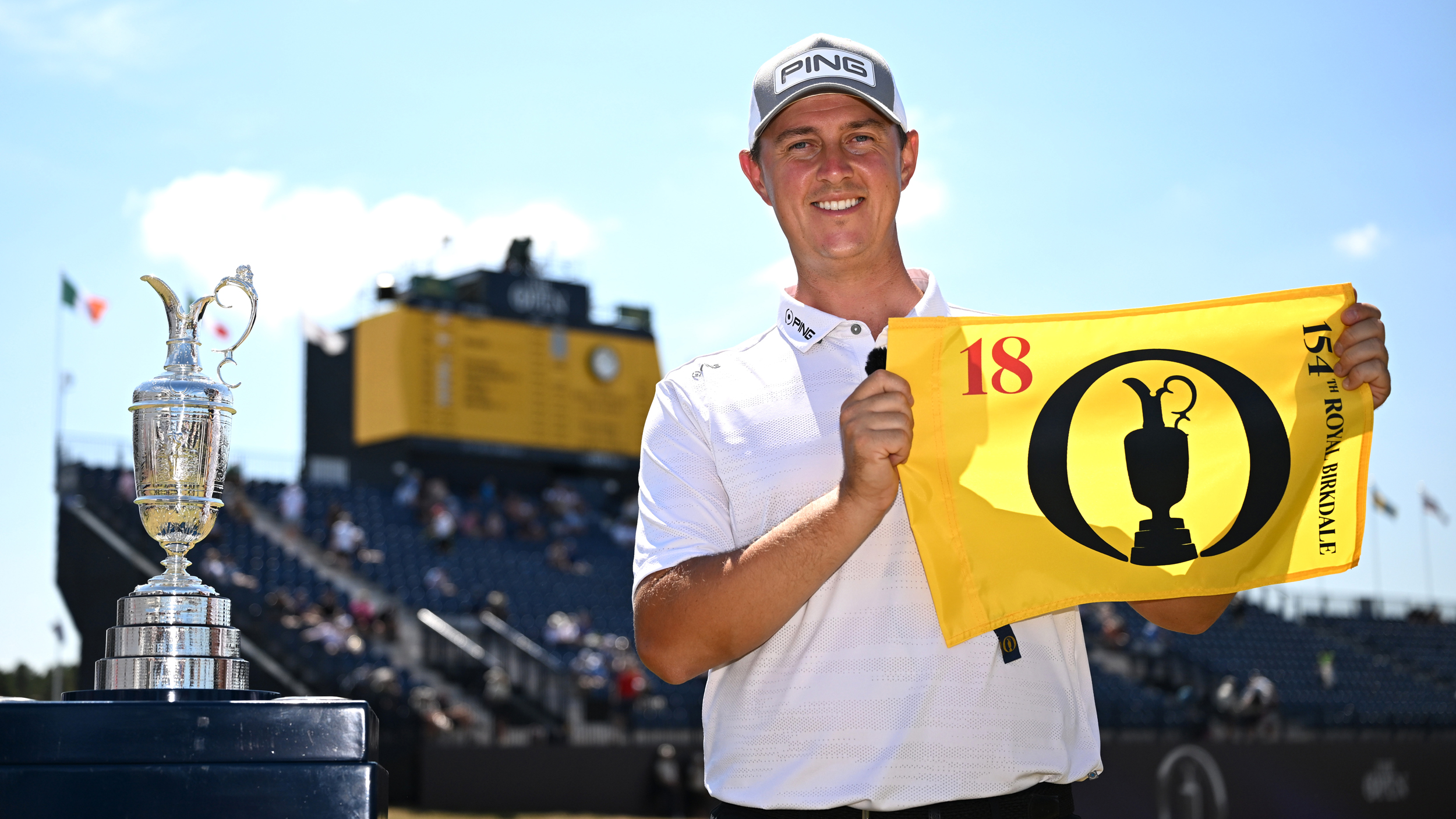 Joe Dean holds a yellow Open flag next to the Claret Jug