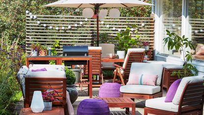 A patio area with wooden furniture, purple pouffes and cushions and white string light decor