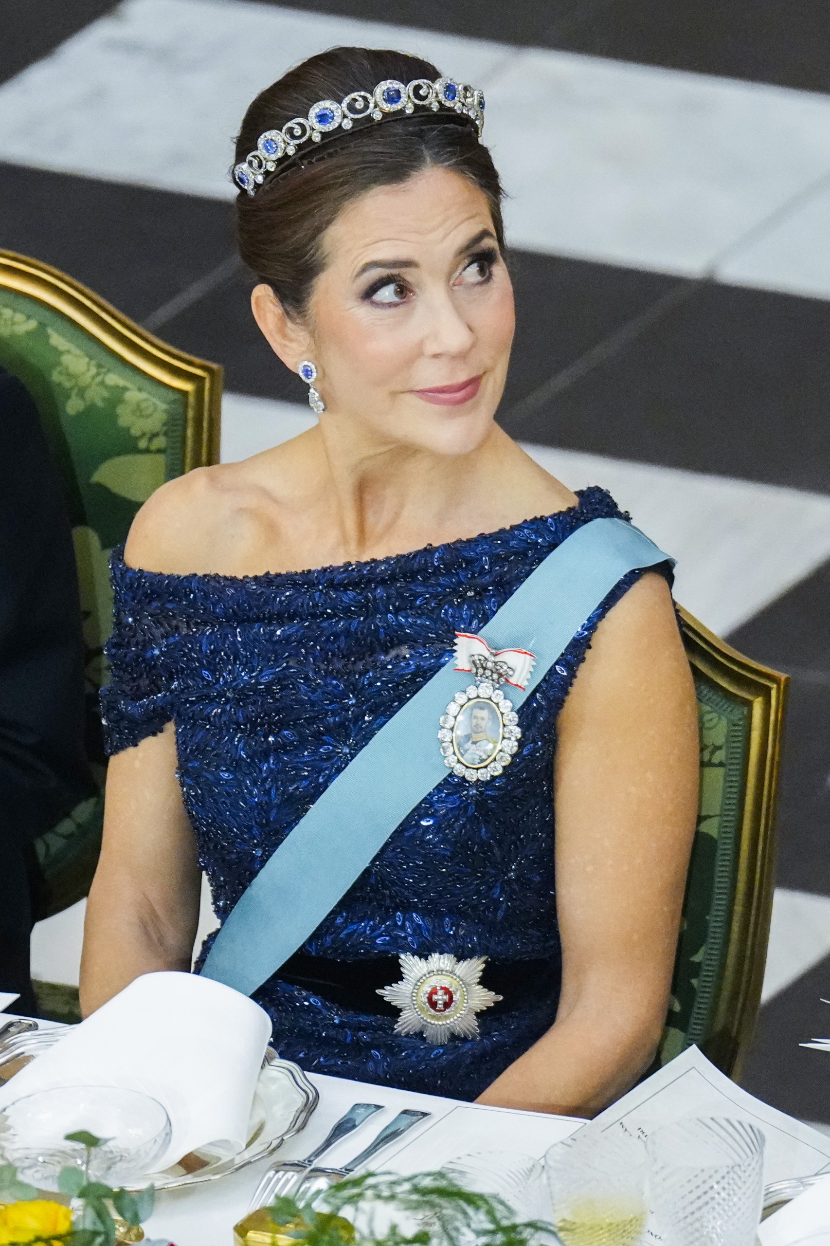 Queen Mary wearing a blue top, sash and tiara sitting at a banquet table