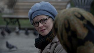 Still from the T.V. show For All Mankind (season 4, episode 1). Close up of 2 women sitting on a park bench. One lady is looking towards us with a concerned look on her face. She is wearing a warm blue hat, black-rimmed glasses, a scarf and a big coat. We can only see the back of the other woman's head, who is wearing a scarf to cover her head. In the background you can see pigeons lurking around another park bench.
