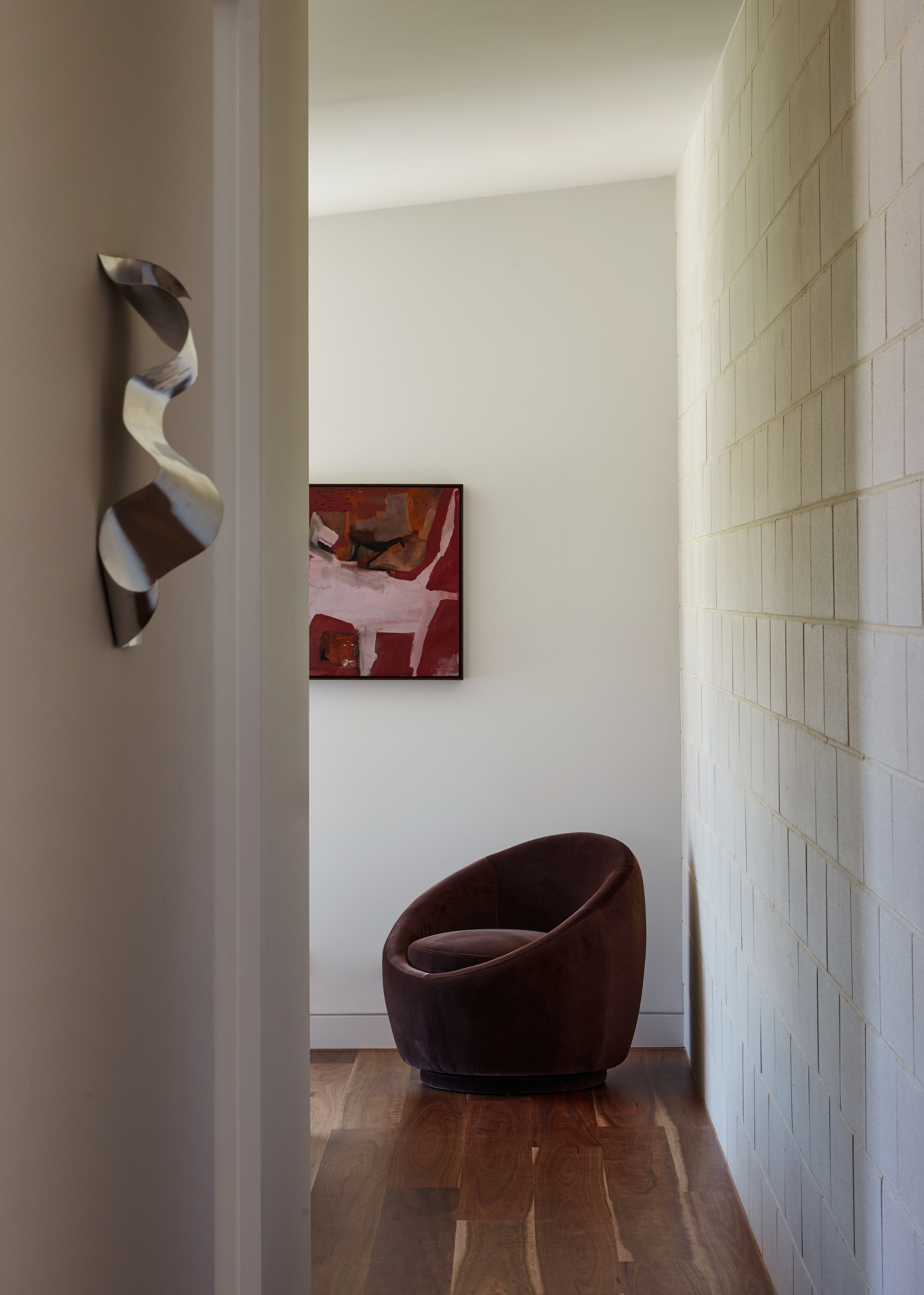 A white hallway with a textured silver wall hanging, a rounded brown chair, and a framed painting