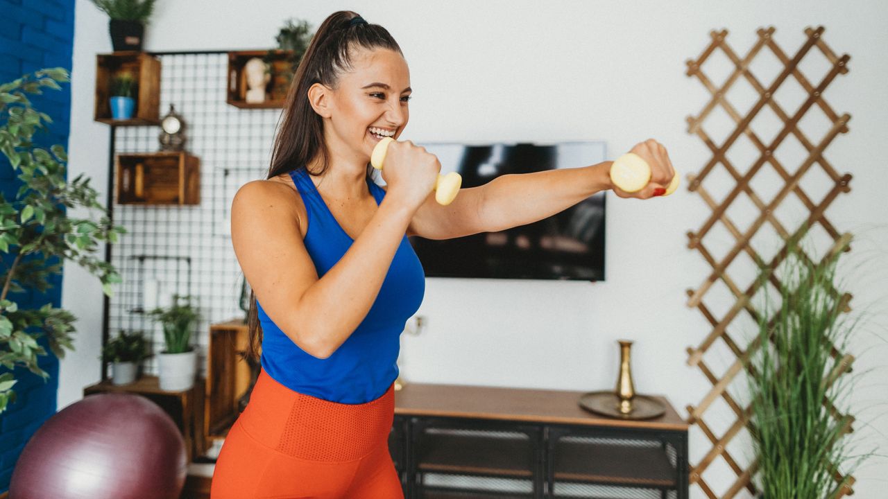 A woman in workout clothes stands smiling and holding light dumbbells in her hand, she is punching forward with her left hand while her right hand rests close to her face. Behind her we see a desk, decorative shelving and a leafy plant.