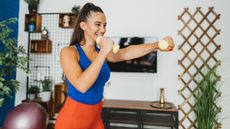 A woman in workout clothes stands smiling and holding light dumbbells in her hand, she is punching forward with her left hand while her right hand rests close to her face. Behind her we see a desk, decorative shelving and a leafy plant.