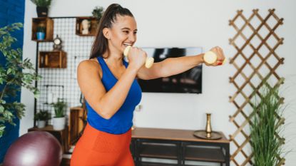 A woman in workout clothes stands smiling and holding light dumbbells in her hand, she is punching forward with her left hand while her right hand rests close to her face. Behind her we see a desk, decorative shelving and a leafy plant.