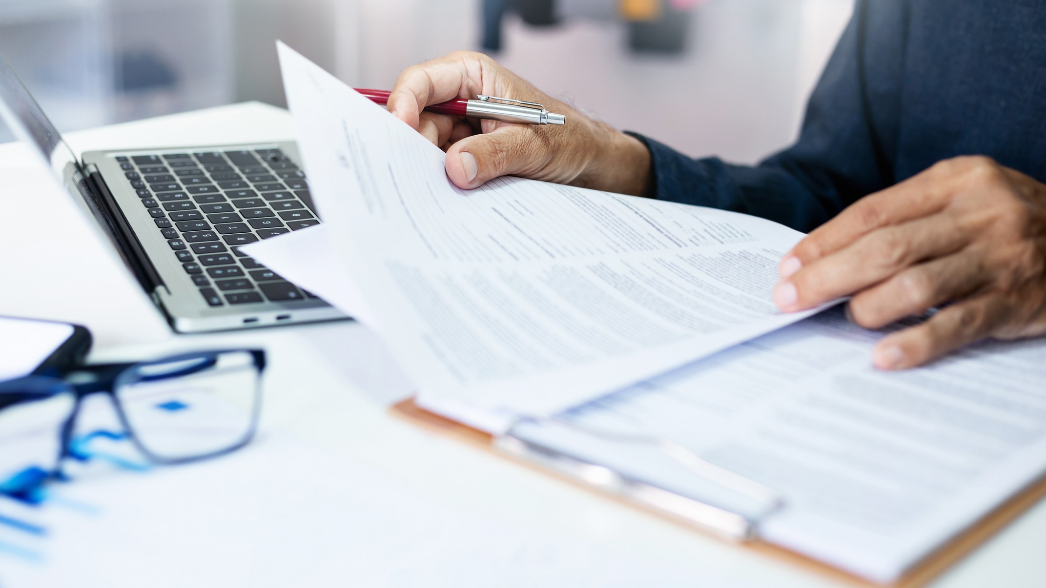 A person is looking through paperwork at a desk with a laptop and reading glasses next to them.