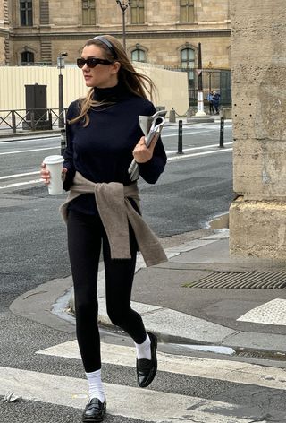 The winter capsule wardrobe item, thermal underpinnings, is shown in a photo of a woman crossing the street wearing a black headband, black sunglasses, a navy turtleneck with a beige sweater tied around the waist styled with thermal leggings, white socks, and black loafers