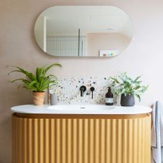 Pink bathroom wall with a lozenge shaped mirror, wooden fluted vanity with white countertop, terrazzo splashback and black tap.