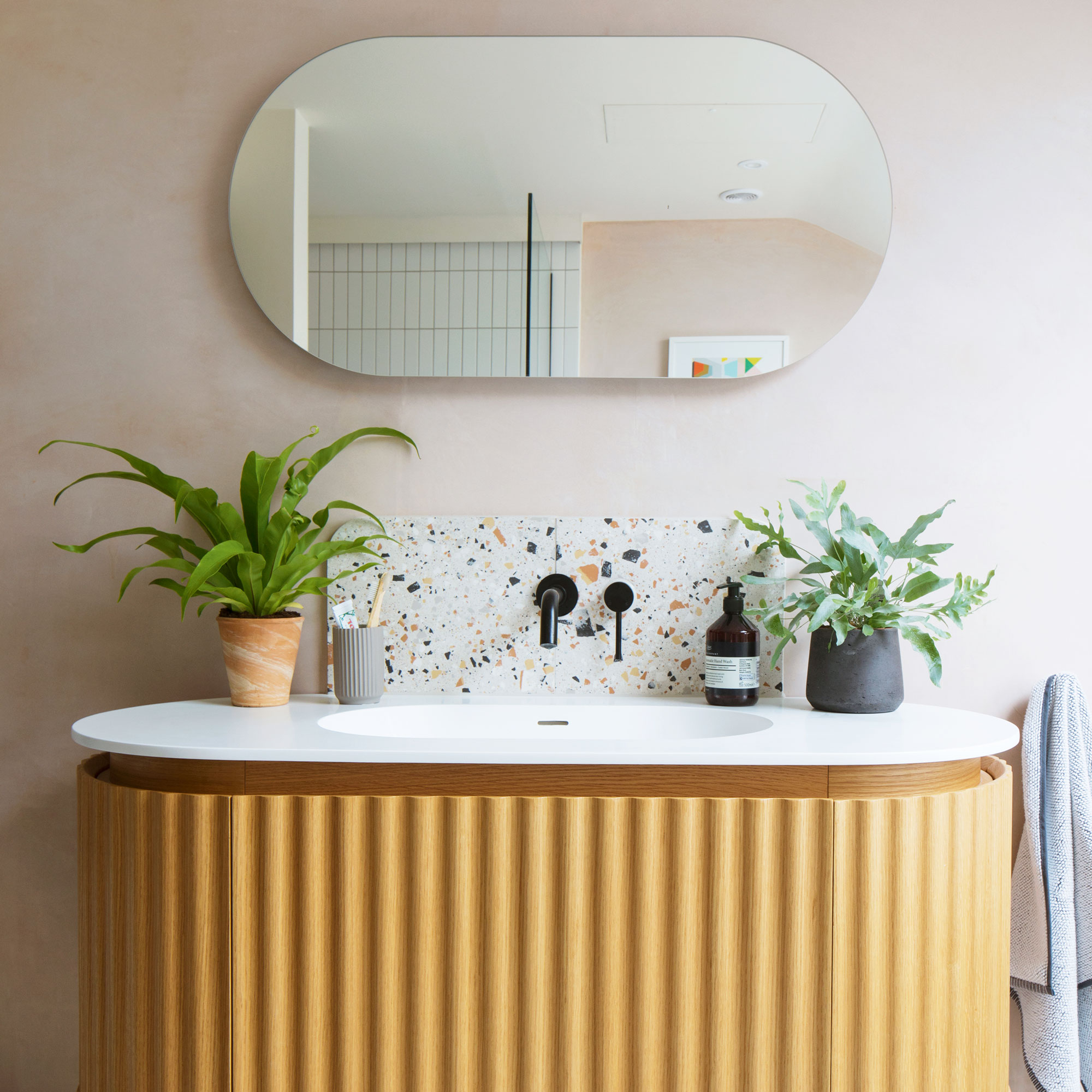 Pink bathroom wall with a lozenge shaped mirror, wooden fluted vanity with white countertop, terrazzo splashback and black tap.