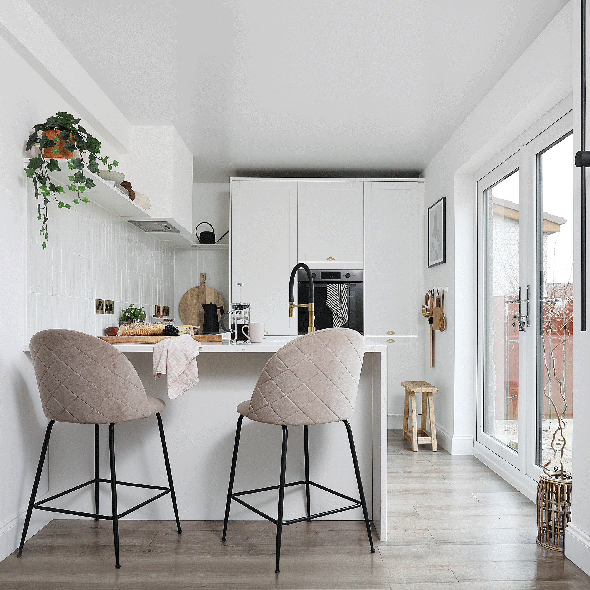 a small white shaker-style kitchen with pale grey wooden flooring and a peninsula breakfast bar with beige quilted upholstered bar stools