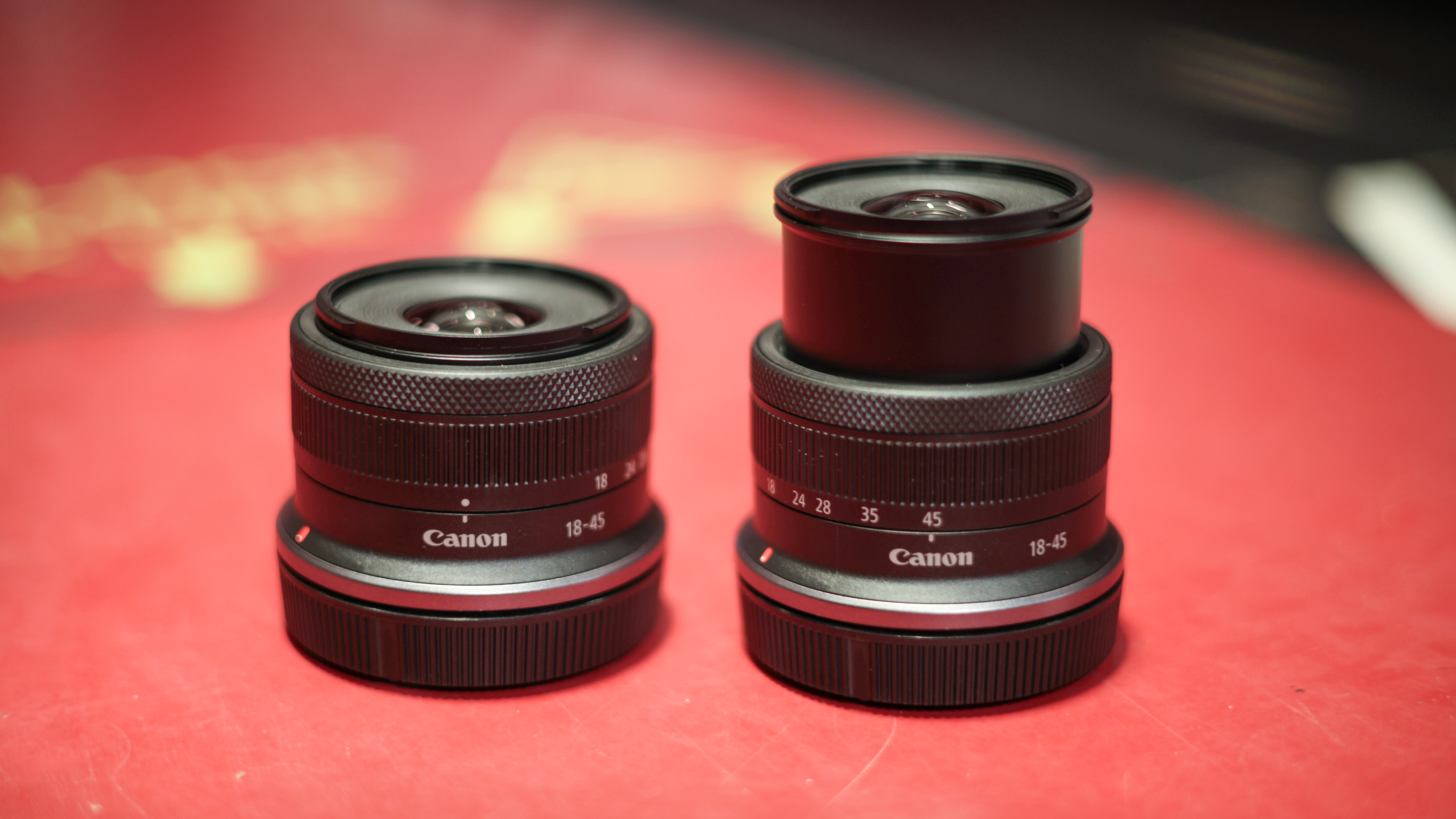 A pair of Canon RF-S 18-45mm f/4.5-6.3 IS STM lenses, one collapsed and one engaged, on a red surface