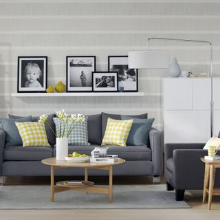 grey living room with a wall-mounted shelf accessorized with picture frames and dark grey sofas with yellow and patterned cushions and a wooden circle shaped coffee table on top of a grey carpet