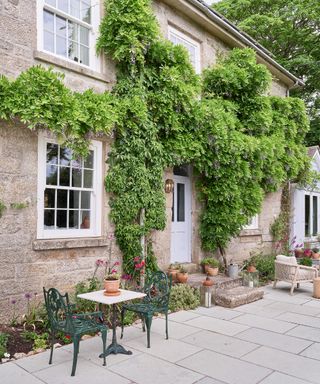 exterior of a Georgian house with wisteria growing up the walls, a pale paved patio and wrought iron garden table and chairs