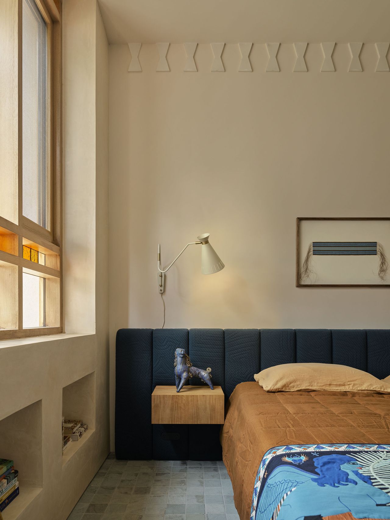 modern bedroom with hempcrete walls and modern wall molding, extended upholstered headboard with orange bedcover and colorful blanket, floating timber side table with statue, wall light, artwork above bed, and timber framed windows with stained glass detail and alcove niches built into wall